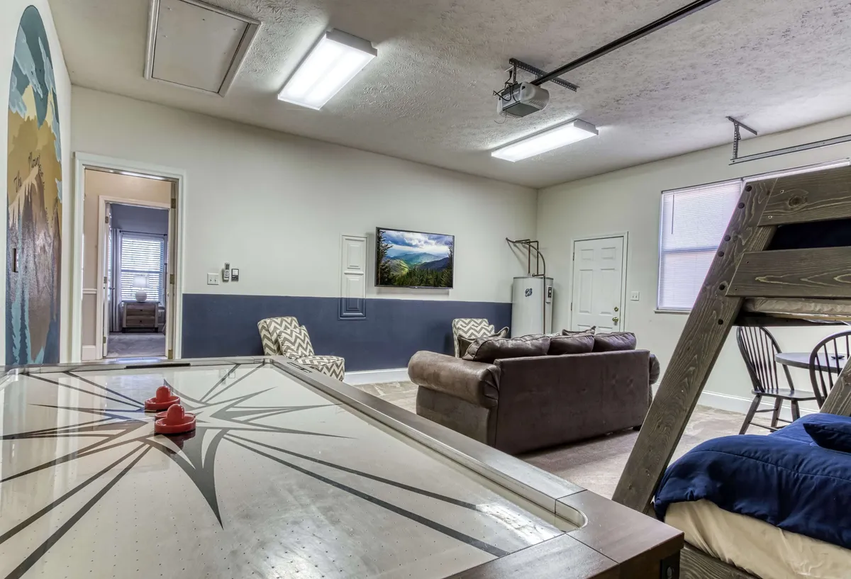 A vibrant, playful game room featuring an air hockey table as the centerpiece, a comfortable sofa seating area, wall mural art, and a television. The space is clearly set up for both gaming and relaxation, encouraging lively gatherings and casual movie nights.