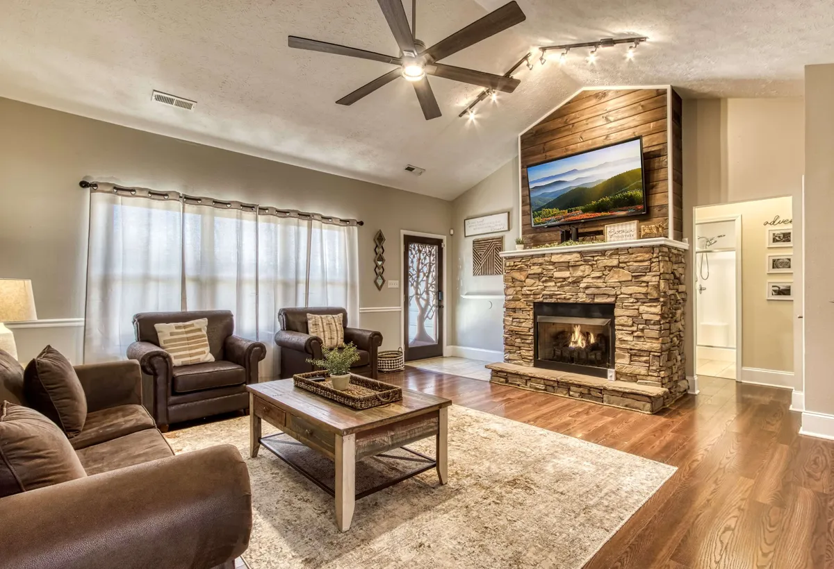 A welcoming living room with a prominent stone fireplace, a large TV, wooden coffee table, and plush seating. The cozy, communal atmosphere is ideal for relaxing, watching movies, or gathering for conversation.