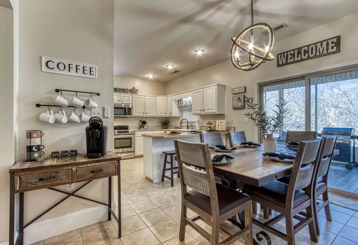 This image features a bright and inviting dining table adjacent to a visible coffee station and modern kitchen appliances. The overall setup suggests a seamless connection between cooking, dining, and socializing, with a welcoming sign and thoughtful decor touches that reinforce a sense of gathering.