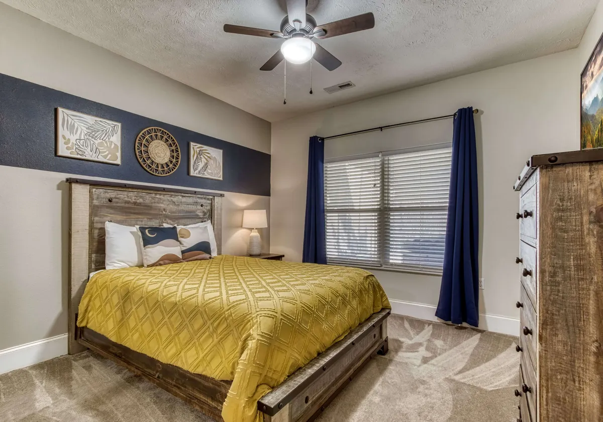 This image features a bed with a cheerful yellow quilt, complemented by wall art, a window with soft curtains, a nightstand with a lamp, and a dresser. The setting exudes a welcoming and tranquil atmosphere ideal for rest and relaxation.