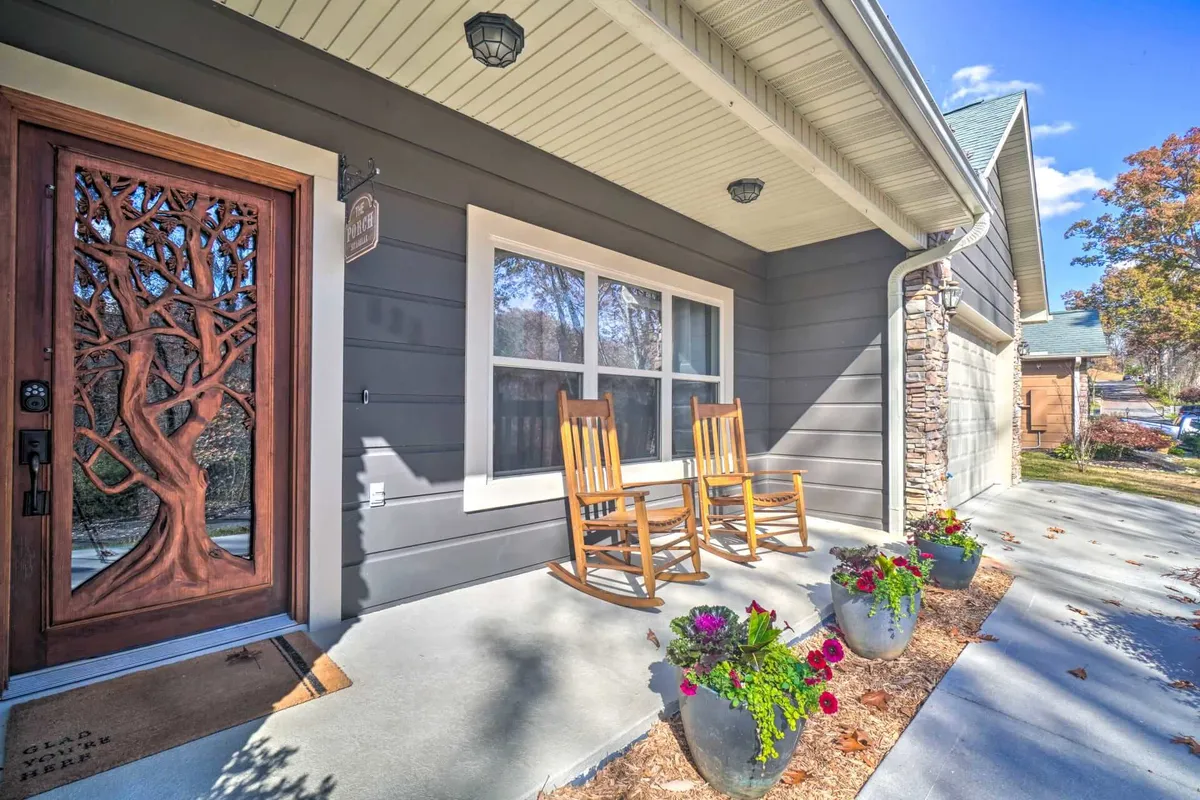 The image displays the inviting front porch of Saddleback Retreat, featuring intricately carved wooden doors, classic rocking chairs, and colorful flower pots brimming with vibrant blooms. The setup captures a welcoming, family-friendly ambiance ideal for relaxing and socializing outdoors.