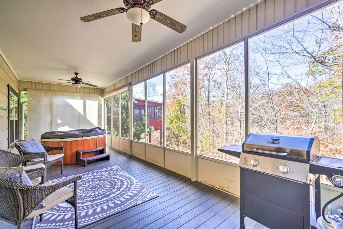This image features a screened-in porch furnished with a hot tub, grill, seating area, and decorative rug, all bathed in natural light from large windows. The space feels comfortable and tranquil, suggesting opportunities for both relaxation and social gatherings.